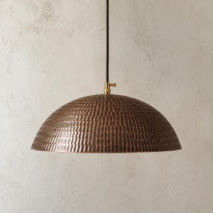 Op de afbeelding: Een koepelvormige hanglamp met een donkerbronskleurige metalen kap met een gehamerd, getextureerd oppervlak. De kap hangt aan een dun zwart snoer en is voorzien van een kleine messingkleurige metalen verbinding aan de bovenzijde.