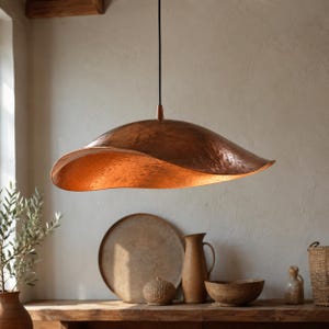 Op de afbeelding: Een hanglamp met een brede, ondiepe koepelvormige kap van gehamerd koper met een golvende, onregelmatige rand. De binnenkant van de metalen kap is verlicht, wat de textuur van het oppervlak en de warme koperen afwerking benadrukt.