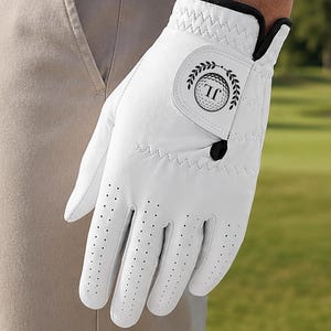 May include: A white golf glove featuring perforated fingers and a black trim. The closure tab displays a circular logo with the letters JL surrounded by a laurel wreath design. The glove is constructed with visible stitching along the wrist.