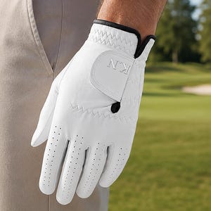 May include: A white golf glove featuring a black trim and a small black tab on the wrist strap. The glove has perforated fingers and the letters KN embossed in silver on the closure flap. The material appears smooth with visible stitching.