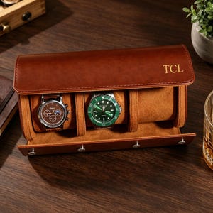 May include: A brown leather watch case with the initials "TCL" embossed in gold. The case is open, revealing two watches with brown and green faces. The case is designed to hold three watches. A glass of amber liquid sits nearby.