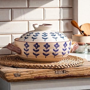 May include: A beige ceramic casserole dish with a lid, adorned with blue floral patterns. The dish has brown handles and sits on a woven mat. The background features white tiles and wooden kitchen utensils.