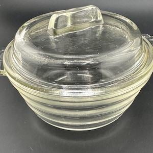 May include: A clear glass casserole dish with a lid. The dish has a ribbed design on the sides and small handles on either side. The lid has a rectangular handle. The dish is sitting on a dark surface.