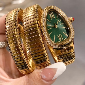 Op de afbeelding: Een goudkleurig horloge met een flexibele, meervoudig gewikkelde band en een druppelvormige kast. Het horloge heeft een diepgroene wijzerplaat met zonnestraalpatroon, gouden Romeinse cijfers op twaalf en zes uur en een lunette bezet met heldere steentjes.
