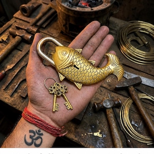 May include: A handcrafted brass padlock in the shape of a fish, held in a hand. The gold-colored lock has intricate scale detailing and comes with two matching keys. The image also shows tools and the text: Handcrafted by Indian Artisans, Traditional Sand Casting Methods Brass Padlock.