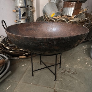 Peut inclure: Un grand récipient de cuisson rond en fer brun foncé avec deux poignées latérales, reposant sur un support en métal noir à quatre pieds fins et une base en croix. La surface du bol présente une patine texturée et usée aux tons sombres.