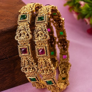 May include: Two gold-toned metal bangles featuring intricate embossed deity figures and alternating square-cut green and pink gemstones. The bangles have a textured, antique-style finish with detailed carvings surrounding each stone setting.