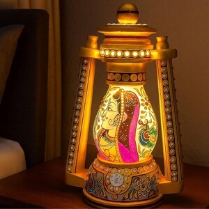Op de afbeelding: Een decoratieve lantaarnvormige lamp met een goudkleurig frame versierd met kleine ingebedde kristallen. Het centrale glazen paneel toont een kleurrijk geschilderd profielportret, geaccentueerd met bloemmotieven en levendige roze tinten.