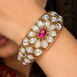 May include: A wide gold-toned cuff bracelet featuring a central oval-shaped deep pink gemstone surrounded by a floral pattern of clear, pear-shaped stones. The metalwork is intricate, with multiple rows of clear stones set in a lattice design.