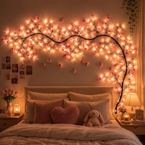 Op de afbeelding: Een wandlamp in de vorm van een uitgestrekte boomtak met talloze kleine, gloeiende roze bloemvormige lampjes. De tak strekt zich horizontaal uit over de muur boven een bed, voorzien van diverse kleine vlindervormige accenten.