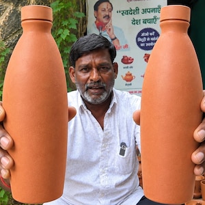 Op de afbeelding: Twee hoge, terracotta-kleurige kleien waterflessen met gladde, ronde lichamen en bijpassende doppen. Elke fles heeft een smalle hals die uitloopt in een bolvormig midden voordat deze licht taps toeloopt naar een platte, stabiele basis.
