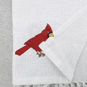 May include: White waffle-weave kitchen towel featuring a detailed illustration of a red cardinal bird. The bird is perched on the towel, with its red feathers and yellow beak clearly visible. The towel has a fringed edge.