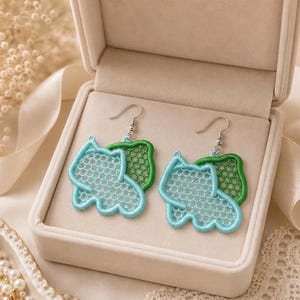 May include: A pair of dangle earrings featuring light blue and green embroidered lace motifs in a scalloped, organic shape. The earrings are attached to silver-toned metal fishhook wires and are presented inside a beige jewelry box.