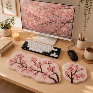 Op de afbeelding: Een bureau-opstelling met een computermonitor met een kersenbloesemafbeelding, een zwart-wit toetsenbord, een zwarte muis en een roze bloemenmuismat en polssteun. Een kopje koffie en een kaars staan ook op het bureau.