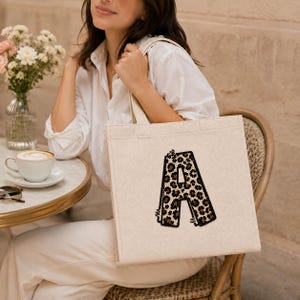 Puede incluir: Bolso tote de lona beige con una gran letra "A" con estampado de leopardo. El bolso tote tiene forma rectangular con un asa sencilla. La letra "A" está delineada en negro.