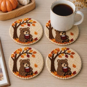 Puede incluir: Cuatro posavasos redondos de ganchillo color crema con un oso pardo sentado bajo un árbol con hojas de otoño naranjas y rojas. Cada oso sostiene una pequeña taza roja. Un posavasos sostiene una taza de cerámica blanca con café.
