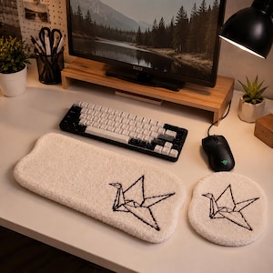 May include: A white tufted keyboard wrist rest and a matching circular mouse pad, both featuring a black embroidered origami crane design. The set is placed on a desk alongside a black mechanical keyboard and a black computer mouse.