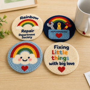 Op de afbeelding: Vier ronde punch needle onderzetters met kleurrijke ontwerpen. Eén is crème met een regenboog en de tekst Rainbow Repair Department Society. Eén is donkerblauw met een gereedschapskist, gereedschap en een rood hart. Eén is lichtblauw met een lachende wolk, regenboog en rood hart. Eén is crème met de tekst Fixing Little things with big love en een rood hart.