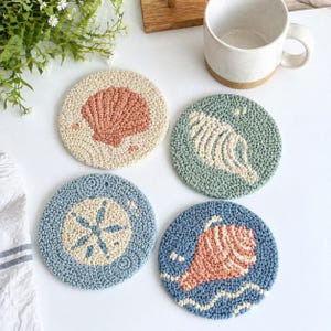 May include: Four circular punch needle coasters featuring coastal motifs. One is cream with a peach seashell, one is sage green with a cream conch shell, one is light blue with a cream sand dollar, and one is blue with a peach conch shell.
