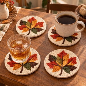 Puede incluir: Un juego de cuatro posavasos redondos de color crema con un diseño de hoja de arce bordado y texturizado en tonos rojo, naranja, amarillo y verde oscuro. Dos posavasos sostienen una taza de café de cerámica y un vaso de té helado.