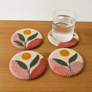 May include: Four round coasters with a floral design. Each coaster features a cream-colored background with a yellow sun, green leaves, and pink and coral-colored petals. A glass of water sits on one of the coasters.