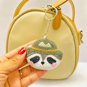 May include: A handmade raccoon keychain with a green hat and a gold-colored band. The keychain is attached to a silver ring. The raccoon has black eyes, a black nose, and pink cheeks. The background includes a light yellow purse.