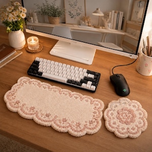 Op de afbeelding: Een rechthoekige getufte polssteun voor een toetsenbord en een bijpassende ronde muismat, beide met een crèmekleurige basis, een geschulpte rand en een delicaat roze bloemenpatroon. De textielaccessoires liggen op een houten bureauoppervlak.