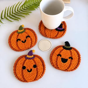 Könnte beinhalten: Drei orangefarbene, kürbisförmige Punch-Needle-Untersetzer mit lächelnden Gesichtern und kleinen Hüten, einer davon unter einer weißen Keramiktasse. Die Untersetzer haben strukturierte Garnschlingen in Orange, Grün, Lila und Schwarz.