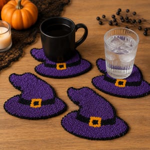 May include: A set of four purple witch hat coasters featuring a black band and a yellow buckle. Each coaster is crafted with a textured punch needle design. The coasters are shaped like pointed hats with a wide brim, providing a decorative surface.
