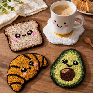 May include: A collection of handmade, textured coasters in the shape of breakfast foods. Includes a slice of toast, a fried egg, a croissant, and an avocado, each with a smiling face. A white mug with coffee sits on the egg coaster.