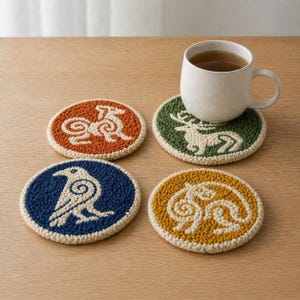 May include: A set of four embroidered round coasters in various colours: orange, green, blue, and yellow. Each coaster features a different animal design, including a deer and a raven. A white mug filled with tea rests on one of the coasters.