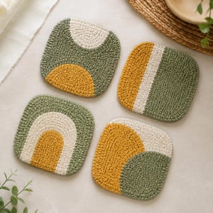 Könnte beinhalten: Ein Set aus vier quadratischen Häkeluntersetzern mit einem strukturierten Strickmuster in Olivgrün, Senfgelb und Creme. Jeder Untersetzer zeigt ein einzigartiges geometrisches Design, darunter Bögen, vertikale Streifen und geschwungene Farbsegmente.