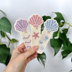 May include: Three handmade crochet bookmarks with seashell designs. Each bookmark features a different color seashell: lavender, pink, and blue. The bookmarks have cream-colored bases with pearl accents and sea-themed embellishments like a starfish and wavy lines.