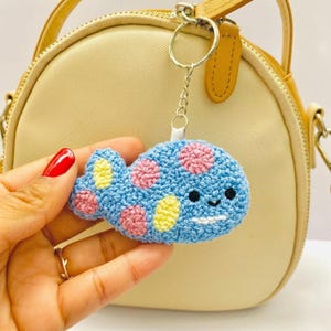 以下が含まれることがあります： ピンクと黄色の水玉模様、黒い刺繍の目、白いステッチの口が特徴の青いかぎ針編みのクジラのキーホルダー。クジラは短いチェーンで銀色の金属製二重リングに取り付けられており、小さなアクセサリーになっています。