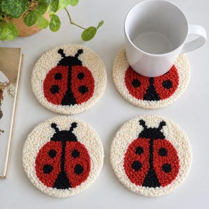 Op de afbeelding: Vier ronde, crèmekleurige onderzetters met een lieveheersbeestjesontwerp. Elke onderzetter heeft een rood lieveheersbeestje met zwarte stippen en een zwarte kop. Een witte mok staat op een van de onderzetters.