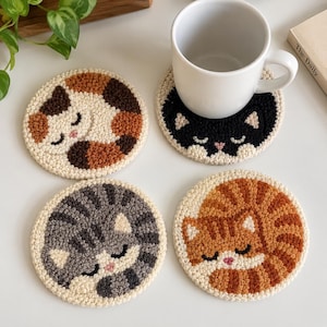 May include: Four circular punch needle coasters featuring sleeping cat designs. One coaster is calico, one is black and white, one is gray tabby, and one is orange tabby. A white ceramic mug sits on the black and white cat coaster.