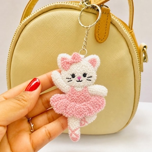 Op de afbeelding: Een witte en roze katten sleutelhanger in een ballerina outfit. De kat heeft een roze strik, een roze tutu en staat op haar tenen. De sleutelhanger is bevestigd aan een zilveren sleutelring. De achtergrond is een lichtgele handtas.