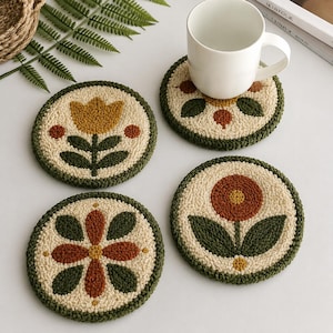 Puede incluir: Cuatro posavasos redondos de punch needle con fondo color crema y bordes verde oscuro. Cada posavasos muestra un motivo floral único en tonos amarillo mostaza, naranja quemado y verde oscuro. Una taza de cerámica blanca descansa sobre uno.