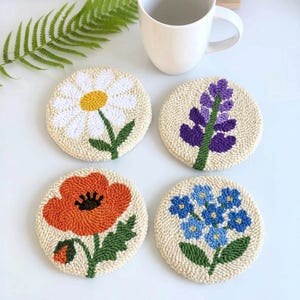 May include: Four round, hand-stitched coasters with floral designs. One features a daisy with white petals and a yellow center. Another has a purple lavender sprig. The third has a red poppy, and the last has blue forget-me-nots. A white mug is in the background.
