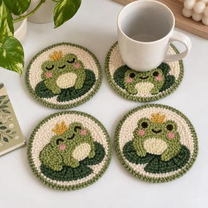 May include: Four circular, hand-tufted coasters featuring a green frog wearing a small yellow crown, sitting on a dark green lily pad against a cream-colored background. The coasters have a textured, looped yarn surface and a green border.