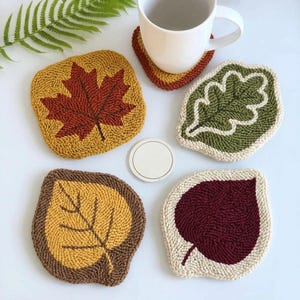 Op de afbeelding: Een verzameling handgemaakte blad-vormige onderzetters in herfstkleuren. De onderzetters hebben esdoornbladeren, eikenbladeren en andere bladeren in bruin-, geel- en groentinten. Een witte mok staat op een bijpassende onderzetter.