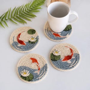 May include: Four round crochet coasters featuring a pond scene with orange koi fish, white water lilies, and green lily pads on a light beige background. One coaster supports a plain white ceramic mug, while the others are arranged nearby.