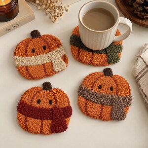 Puede incluir: Cuatro posavasos redondos con forma de calabaza hechos de hilo naranja texturizado. Cada calabaza tiene un pequeño tallo marrón oscuro, dos ojos negros y una bufanda envuelta en color crema, verde oscuro, rojo oscuro o marrón. Una taza blanca descansa sobre uno.