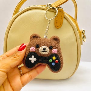 May include: A brown, crocheted bear-shaped keychain with a black game controller design. The controller features a white cross, colorful buttons, and a small, round face. The keychain is attached to a silver ring.