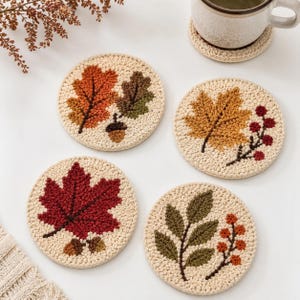 May include: Four round, cream-colored crocheted coasters featuring textured autumn-themed designs. Each coaster displays different motifs including orange and brown oak leaves with an acorn, a yellow maple leaf with red berries, a red maple leaf, and green leaves with orange berries.