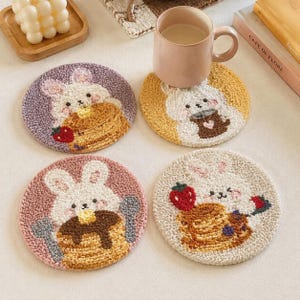 May include: Four round punch needle coasters featuring white bunny illustrations. One shows a bunny with pancakes and a strawberry, another with a mug, one with pancakes and utensils, and the last with pancakes and berries. Each has a unique background color.