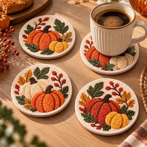 Puede incluir: Un juego de cuatro posavasos redondos con diseños texturizados de calabazas naranjas y amarillas, hojas verdes y bayas rojas sobre un fondo color crema. Un posavasos sostiene una taza de cerámica blanca llena de café negro.