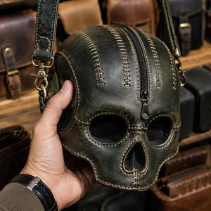 May include: A dark olive green leather bag shaped like a human skull. The bag features visible light-coloured stitching along the seams and a central black zipper closure running vertically from the top to the nasal cavity area.