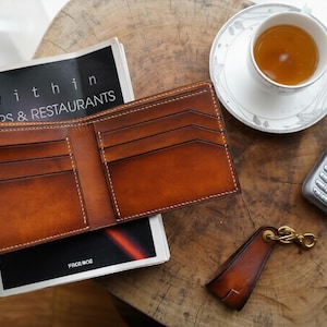 May include: Open brown leather wallet with multiple card slots, resting on a magazine with the text "within BARS & RESTAURANTS". A cup of tea, a silver flip phone, and a leather keychain are also on the wooden surface.