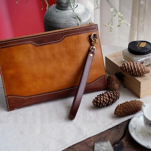 Puede incluir: Clutch de cuero marrón con ribete marrón oscuro y costuras blancas. Incluye una correa de muñeca y herrajes dorados. El clutch es rectangular y tiene cierre de cremallera.
